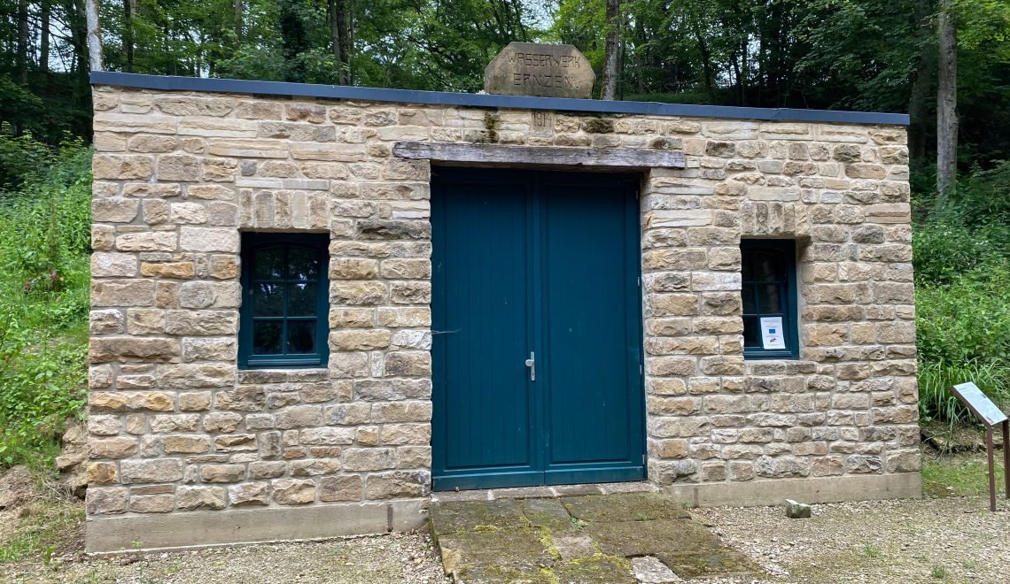 Pumpenhaus aus Sandstein mit grüner Doppeltür und dem Schriftstein „Wasserwerk Ernzen“